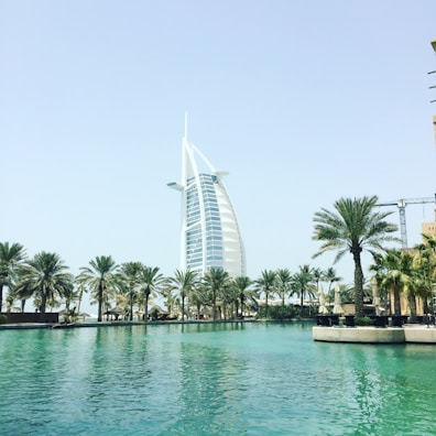 A large sail-shaped building, surrounded by palm trees, stands beside a tranquil body of water on a bright, sunny day. The structure is well-known for its modern and unique architectural design.