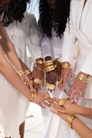 A group of individuals with diverse skin tones gather in a circle, extending their hands adorned with matching gold jewelry, including ornate bracelets and rings. They are wearing elegant white clothing, symbolizing unity and harmony.