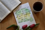 An open book is placed next to a handwritten note in German along with a teacup and two red tulips. The note has artistic watercolor splashes in green and yellow.