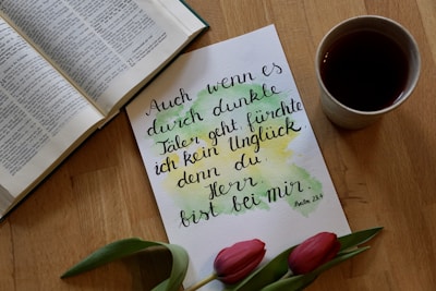 An open book is placed next to a handwritten note in German along with a teacup and two red tulips. The note has artistic watercolor splashes in green and yellow.