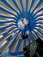 A symmetrical, circular architectural structure featuring a combination of glass and steel elements radiating from the center. The intricate design resembles a fan or a spider web with alternating bands of transparent panels and cables connecting to a central hub.
