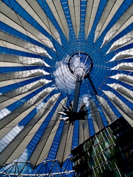 A symmetrical, circular architectural structure featuring a combination of glass and steel elements radiating from the center. The intricate design resembles a fan or a spider web with alternating bands of transparent panels and cables connecting to a central hub.