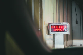 A digital sign with bright red LED text displays the words 'HIGH IMPACT' mounted on a wall in an industrial or warehouse setting. The environment features vertical metal panels and subdued lighting.