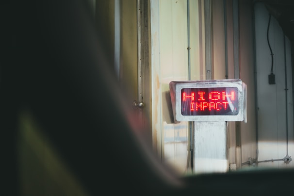 A digital sign with bright red LED text displays the words 'HIGH IMPACT' mounted on a wall in an industrial or warehouse setting. The environment features vertical metal panels and subdued lighting.