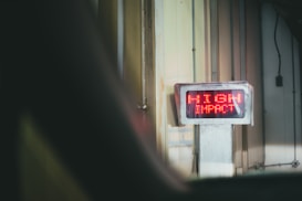 A digital sign with bright red LED text displays the words 'HIGH IMPACT' mounted on a wall in an industrial or warehouse setting. The environment features vertical metal panels and subdued lighting.