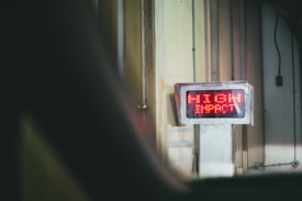 A digital sign with bright red LED text displays the words 'HIGH IMPACT' mounted on a wall in an industrial or warehouse setting. The environment features vertical metal panels and subdued lighting.