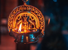 An intricately designed brass oil lamp with a vivid flame is depicted. The lamp features detailed carvings and appears to resemble a traditional Indian design or Hindu iconography. The warm glow from the flame illuminates the rich golden hues of the lamp, creating a serene and contemplative atmosphere.