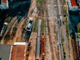 An aerial view of a railway yard featuring multiple train tracks diverging and converging. A train is positioned on one of the tracks alongside various industrial buildings and equipment. The area surrounding the tracks has a mixture of grass and gravel, with some old structures scattered throughout.