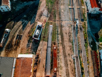 An aerial view of a railway yard featuring multiple train tracks diverging and converging. A train is positioned on one of the tracks alongside various industrial buildings and equipment. The area surrounding the tracks has a mixture of grass and gravel, with some old structures scattered throughout.