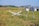 A small white aircraft is crashed into a field of grass next to an old metal swingset. In the background, there are several small buildings with one appearing to be a barn or shed. The landscape is rural with rolling hills and a few scattered trees.