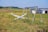 A small white aircraft is crashed into a field of grass next to an old metal swingset. In the background, there are several small buildings with one appearing to be a barn or shed. The landscape is rural with rolling hills and a few scattered trees.