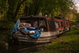 A narrowboat is moored alongside a calm, forested riverbank. The vessel is covered with various tarps and ropes, suggesting it's used for long-term living or travel. Surrounded by lush green trees, the boat is adorned with items like a pot, bucket, and other personal effects, conveying a cozy and lived-in feeling.