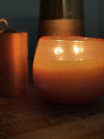 A warm, softly lit scene featuring a glowing candle encased in an orange and yellow gradient glass holder. The candle emits a gentle glow, casting shadows on the surrounding objects, including another block-shaped candle on the left. The muted background enhances the warm, cozy ambiance.