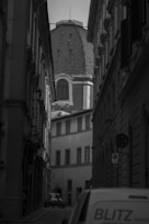 A narrow European street lined with tall, historic buildings leads towards a large, old dome in the background. The scene is captured in black and white, creating a timeless and classic atmosphere. A sign with the word 'hotel' can be seen on the left, and a partial view of a vehicle with the text 'BLITZ' is visible in the foreground.