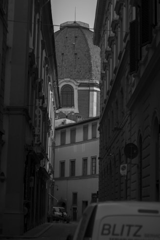 A narrow European street lined with tall, historic buildings leads towards a large, old dome in the background. The scene is captured in black and white, creating a timeless and classic atmosphere. A sign with the word 'hotel' can be seen on the left, and a partial view of a vehicle with the text 'BLITZ' is visible in the foreground.