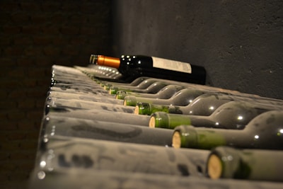 A row of dusty wine bottles is neatly arranged on a cellar rack. The bottles have a green tint and are corked, with one prominent bottle featuring a dark label and orange-tinted capsule standing out among the others. The background shows a dimly lit brick wall, creating a rustic and vintage atmosphere.