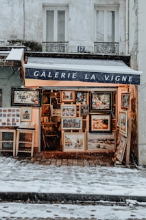 A quaint art gallery storefront with a blue awning and the name 'Galerie La Vigne' prominently displayed. The exterior is adorned with a variety of framed paintings and photographs, creating a warm glow against the cold, snowy environment. Snow is gently falling and accumulating on the cobblestone path in front of the gallery.