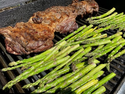 A grill features a large, seasoned piece of meat on the left side, alongside several fresh asparagus spears on the right. The meat appears to be well-seared with a charred surface. The asparagus is vibrant green, freshly cooked, and placed directly on the grill grates.