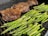 A grill features a large, seasoned piece of meat on the left side, alongside several fresh asparagus spears on the right. The meat appears to be well-seared with a charred surface. The asparagus is vibrant green, freshly cooked, and placed directly on the grill grates.