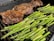 A grill features a large, seasoned piece of meat on the left side, alongside several fresh asparagus spears on the right. The meat appears to be well-seared with a charred surface. The asparagus is vibrant green, freshly cooked, and placed directly on the grill grates.