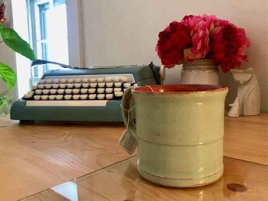 A vintage typewriter sits on a wooden desk beside a mug and a vase filled with vibrant red flowers. A small decorative object resembling a white figure adds an artistic touch to the setting. The ambiance suggests a blend of nostalgia and warmth, enhanced by natural light streaming through a nearby window.