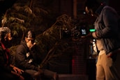 Three individuals are engaged in a filming setup, with one person operating a camera while the other two are seated and appear to be in conversation. The scene is dimly lit and surrounded by greenery, creating a moody atmosphere.