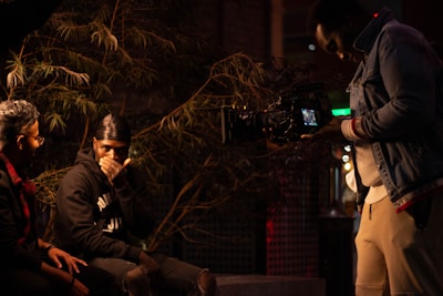 Three individuals are engaged in a filming setup, with one person operating a camera while the other two are seated and appear to be in conversation. The scene is dimly lit and surrounded by greenery, creating a moody atmosphere.