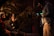 Three individuals are engaged in a filming setup, with one person operating a camera while the other two are seated and appear to be in conversation. The scene is dimly lit and surrounded by greenery, creating a moody atmosphere.