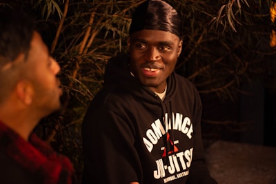 Two people are sitting together in a dimly lit outdoor setting, engaged in conversation. One person, wearing a black hoodie with 'Dominance Jiu-Jitsu' written on it, is smiling warmly. The background is filled with foliage and shadows, suggesting it might be nighttime.