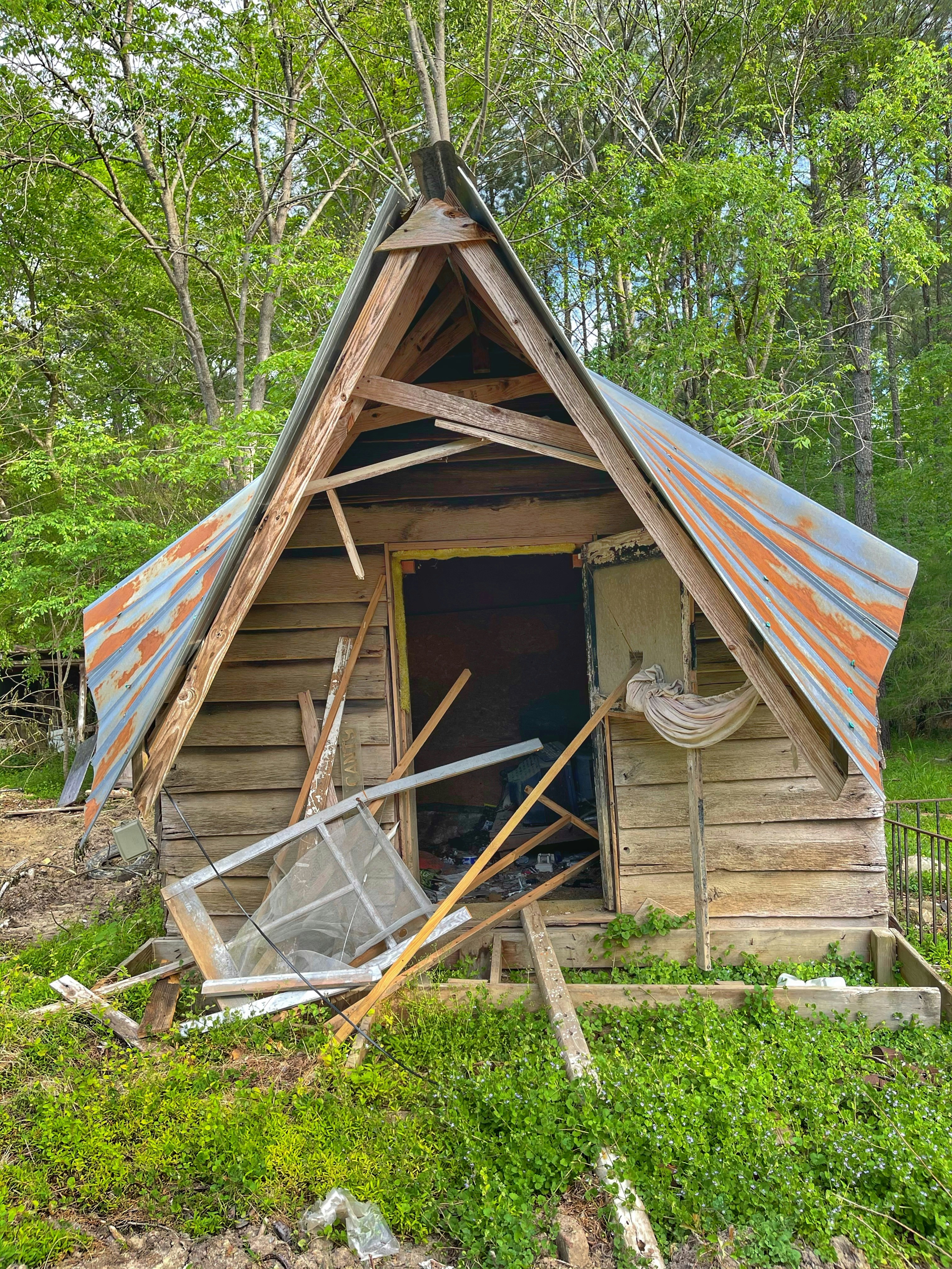 Old rotting wooden shed in a backyard with peeling paint and collapsed roof, overgrown weeds surrounding it
