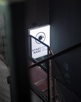 A dimly lit industrial stairwell with a metal mesh railing is partially illuminated by a glowing rectangular sign. The sign has a minimalist design featuring a circle with an orbit-like line and the words 'START BASE NONOS STORY INSPIRA' in a contemporary font.