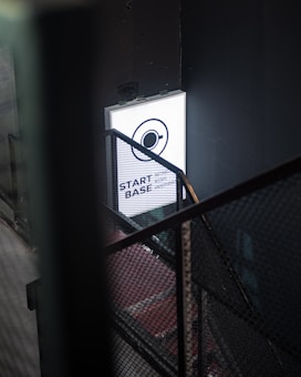 A dimly lit industrial stairwell with a metal mesh railing is partially illuminated by a glowing rectangular sign. The sign has a minimalist design featuring a circle with an orbit-like line and the words 'START BASE NONOS STORY INSPIRA' in a contemporary font.