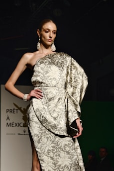 A model is wearing an elegant, patterned gown with one shoulder exposed, adorned with large earrings and a bracelet. The background is dark, and there is a sign with French text partially visible.