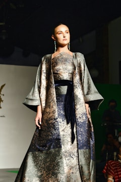A woman is walking down a fashion runway, wearing an elegant, flowing outfit with a metallic and patterned design. The setting appears to be a fashion show with a dark background and stage lighting focused on the model.