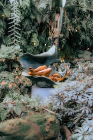 A serene garden scene featuring a bronze sculpture of a reclining figure wrapped in a large leaf. The environment is lush with various green plants and ferns, and small orange flowers add pops of color. Soft ambient lighting creates a peaceful and tranquil ambiance.