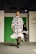 A fashion model walks down a runway in a high-fashion ensemble featuring a white dress with bold black geometric patterns and ruffled details. The background is a bright green panel flanked by white walls, with text and logos visible. Another model is partially visible in the background.