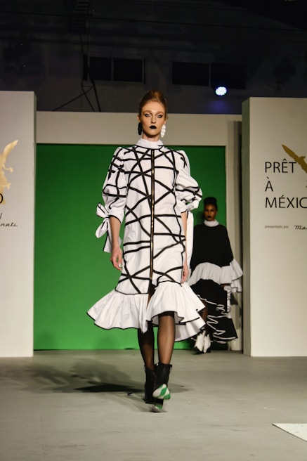 A fashion model walks down a runway in a high-fashion ensemble featuring a white dress with bold black geometric patterns and ruffled details. The background is a bright green panel flanked by white walls, with text and logos visible. Another model is partially visible in the background.
