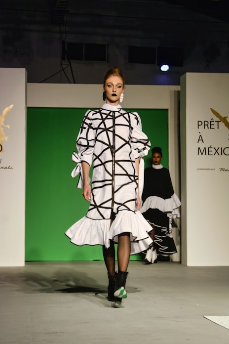 A fashion model walks down a runway in a high-fashion ensemble featuring a white dress with bold black geometric patterns and ruffled details. The background is a bright green panel flanked by white walls, with text and logos visible. Another model is partially visible in the background.