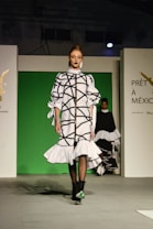 A fashion model walks down a runway in a high-fashion ensemble featuring a white dress with bold black geometric patterns and ruffled details. The background is a bright green panel flanked by white walls, with text and logos visible. Another model is partially visible in the background.