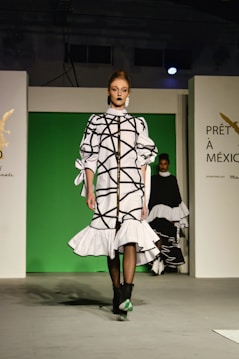 A fashion model walks down a runway in a high-fashion ensemble featuring a white dress with bold black geometric patterns and ruffled details. The background is a bright green panel flanked by white walls, with text and logos visible. Another model is partially visible in the background.