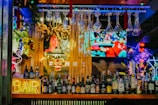 An illuminated bar counter features a variety of liquor bottles, with colorful neon lights adding vibrancy. Glasses hang upside down above the bar, and a large television screen in the background displays a sports event.