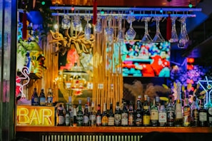 An illuminated bar counter features a variety of liquor bottles, with colorful neon lights adding vibrancy. Glasses hang upside down above the bar, and a large television screen in the background displays a sports event.