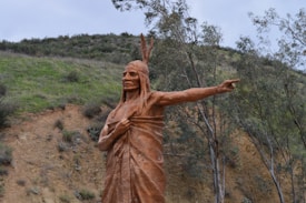 A large statue resembling a Native American figure stands in a natural setting. The figure is carved in a reddish-brown material, and is depicted with traditional headdress and attire. It points dramatically toward the horizon. Surrounding the statue are rolling hills covered with grass and sparse trees, under an overcast sky.