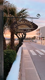 A wooden sign with the text 'El Bungalow Lubina a la Sal' is mounted on a decorative metal bracket, hanging in front of a row of palm trees. The street is lined with a low white wall, leading to a sidewalk and a road with a pedestrian crossing. In the background, there are residential buildings under a serene sky with hues of pink and blue.