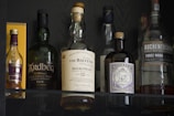 Several bottles of alcohol are displayed on a shelf, including various brands of whisky and gin. The bottles have labels with distinct designs and colors, and they are positioned in a way that showcases their branding. The lighting highlights the reflective surfaces of the bottles.