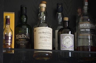 Several bottles of alcohol are displayed on a shelf, including various brands of whisky and gin. The bottles have labels with distinct designs and colors, and they are positioned in a way that showcases their branding. The lighting highlights the reflective surfaces of the bottles.