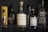Several bottles of alcohol are displayed on a shelf, including various brands of whisky and gin. The bottles have labels with distinct designs and colors, and they are positioned in a way that showcases their branding. The lighting highlights the reflective surfaces of the bottles.