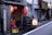 An urban street scene outside a small, traditional Japanese izakaya with several red paper lanterns hanging near the entrance. A bicycle is propped up against a lamppost next to a potted plant and an illuminated sign displaying 'Kirin' in red letters. A bright orange traffic cone is on top of a yellow crate near a metal utility door.