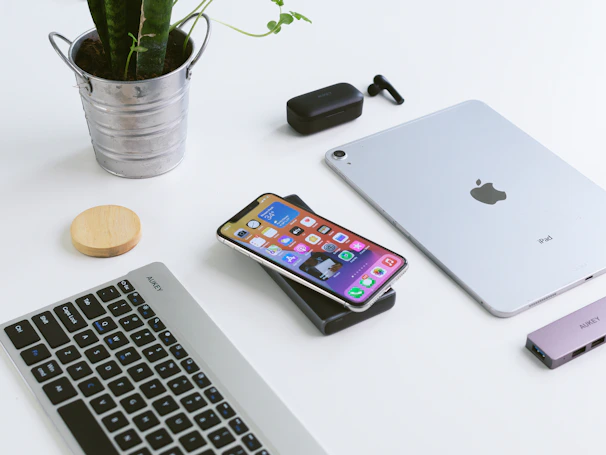 A minimalist workspace arrangement on a white surface includes a silver iPad, a smartphone with colorful apps displayed, a wireless keyboard, a small silver pail containing a plant, wireless earbuds, and a small docking device.