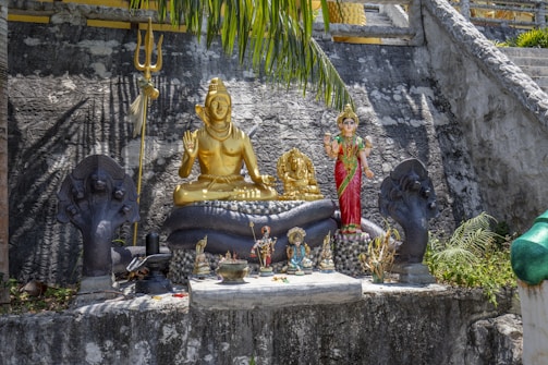 A serene outdoor shrine featuring a golden statue of a deity sitting cross-legged with multiple arms, alongside various other figurines. A trident is positioned nearby, and there are statues of cobras with raised hoods flanking the central figures. The setting includes stone steps and lush greenery, creating a peaceful and spiritual atmosphere.